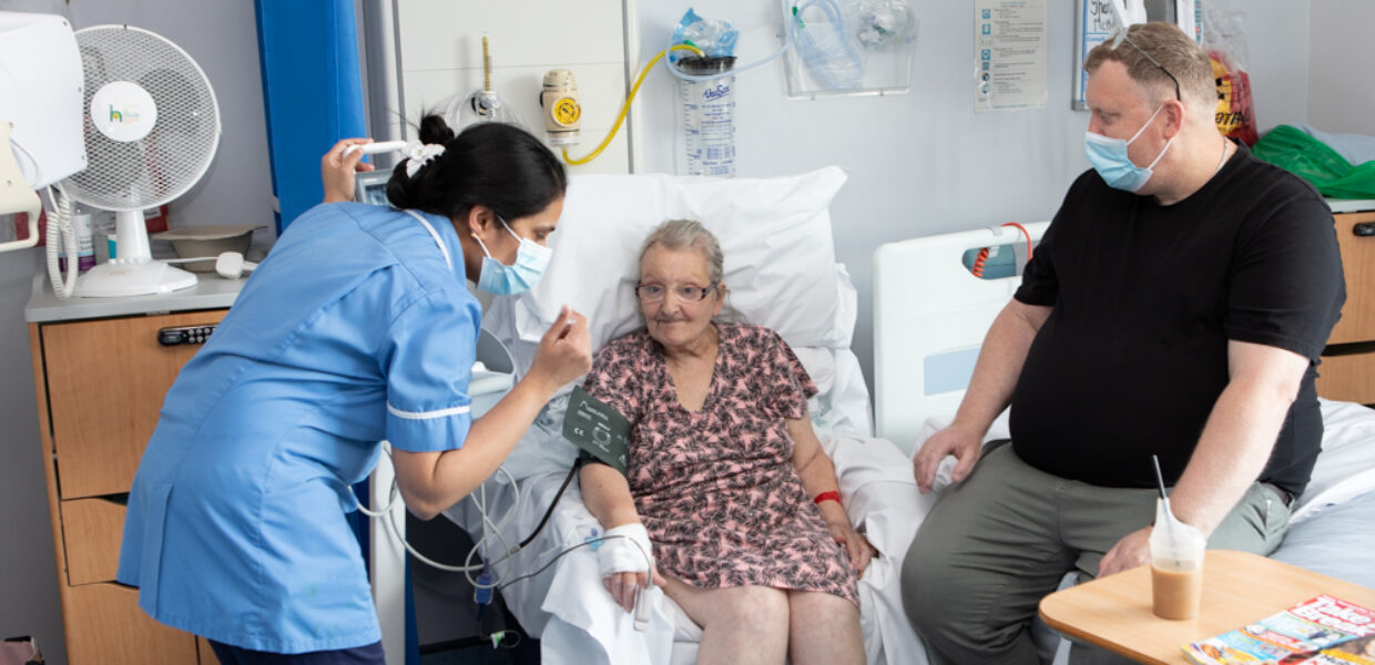 A man visiting his wife sits on the bed and looks on, as she is tended to by a nurse.