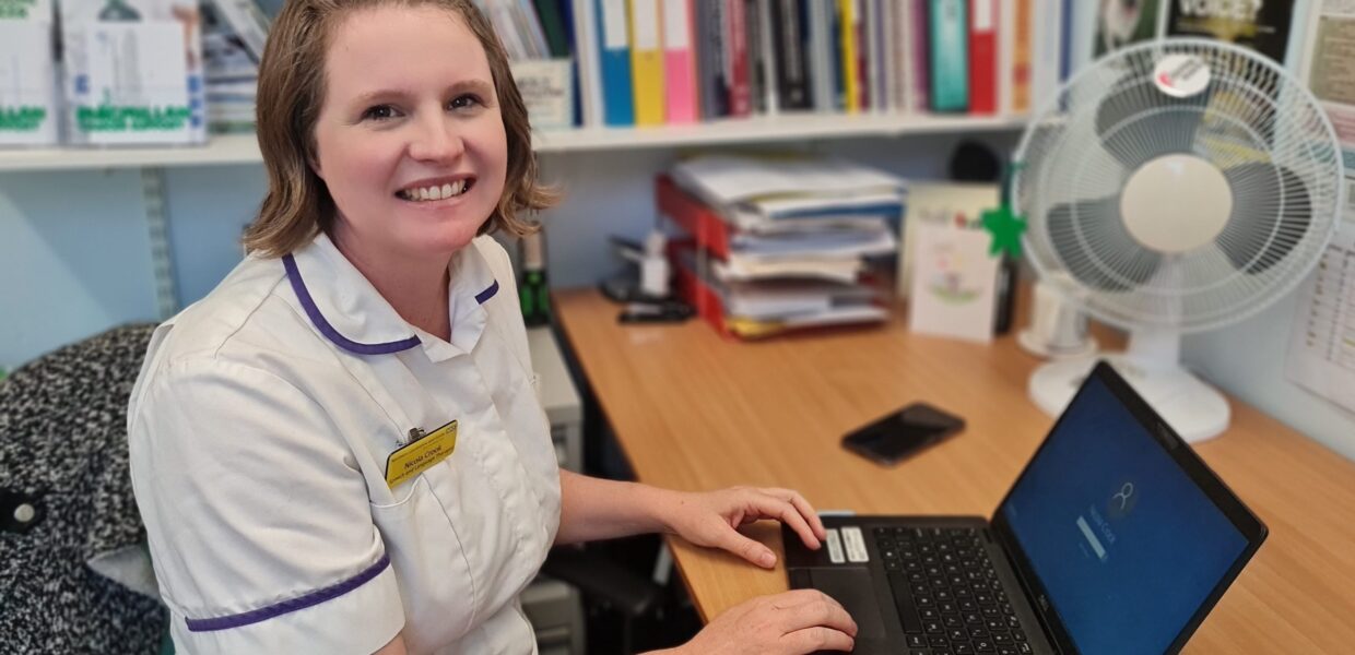 Specialist Speech and Language Therapist (SLT) Dr Nicola Crook led Neil's therapy following his severe stoke. She is seated at a desk in her clinic. In the background are book shelves and a laptop is on the desk in front of her.