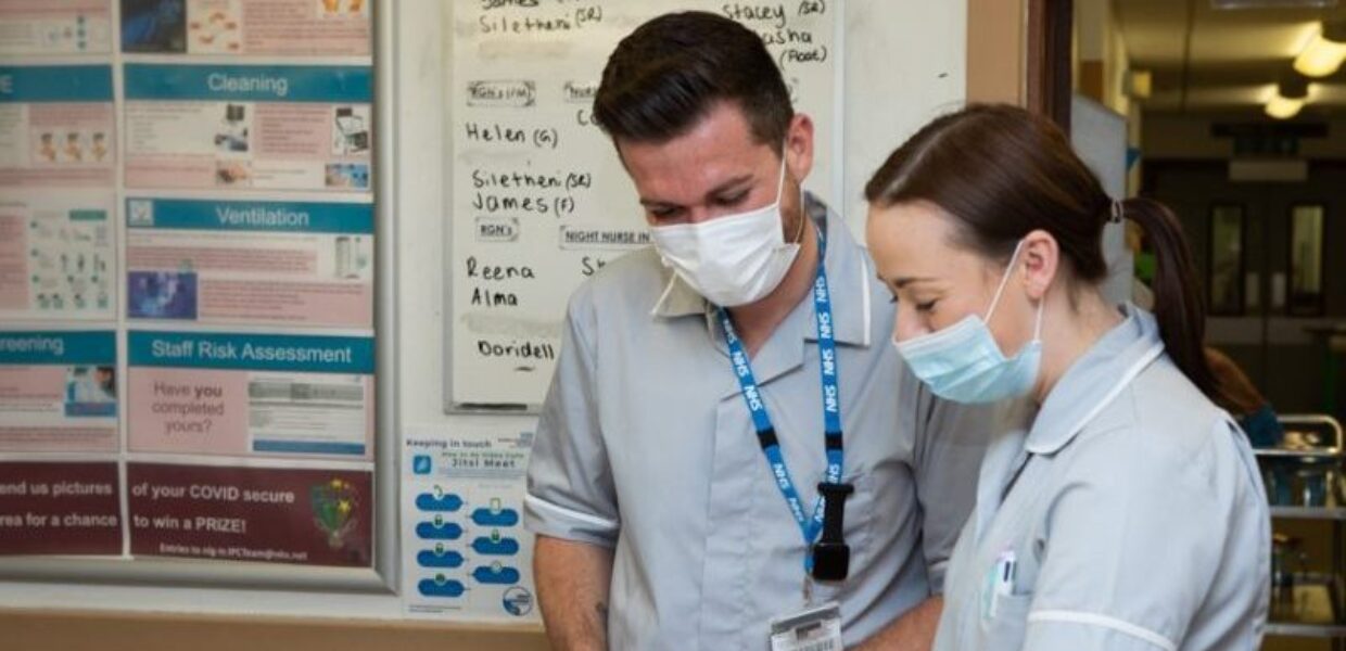 Two healthcare assistants stand at a nursing station looking down at paperwork