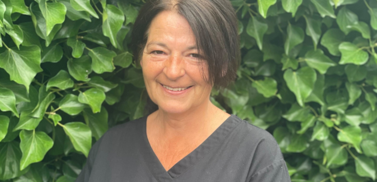 Lynn Sherlock stands smiling outside with greenery in the background, wearing a black tunic