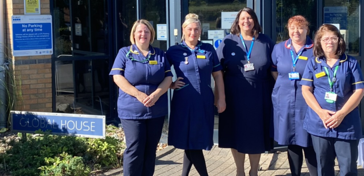 Five members of senior nursing staff stand together outside in their dark blue nursing tunics