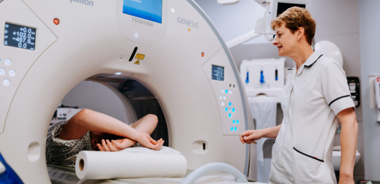 a patient laid down with their arms above their head inside a scanner with a member of staff watching on
