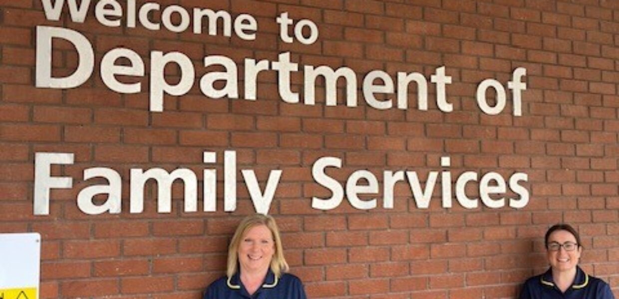 two midwives in blue uniform stood next to a brick wall with a sign reading 'welcome to the department of family services