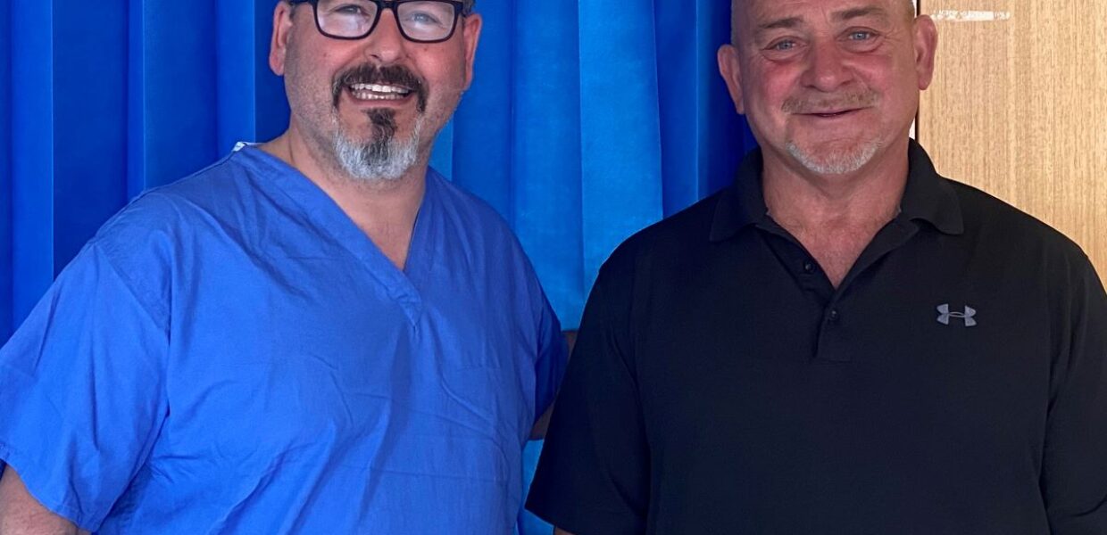 Two men, one dressed in blue theatre scrubs uniform, are standing next to each other